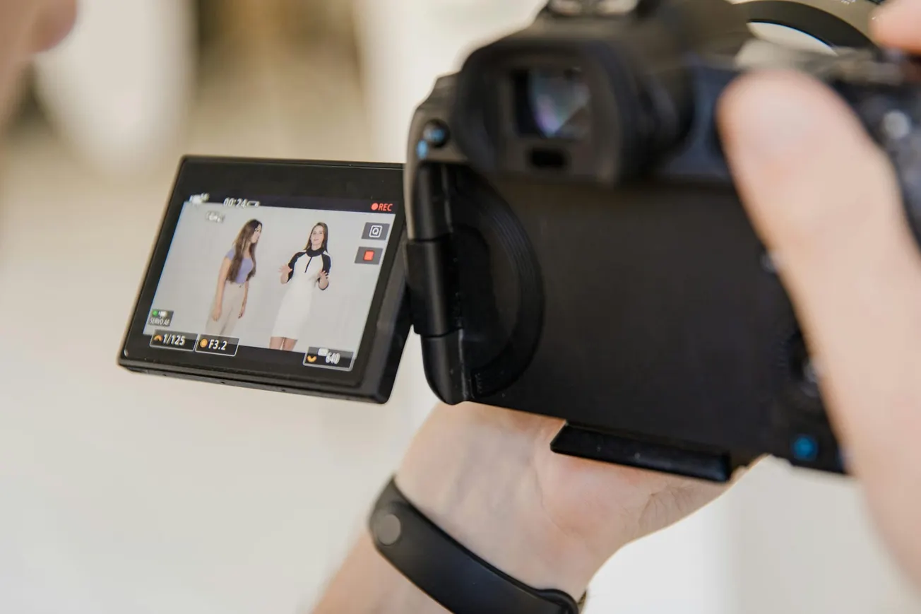 Close-up of a camera screen showing two women in dresses, one holding a microphone. The camera's held by a person with a black wristband, focused on filming.