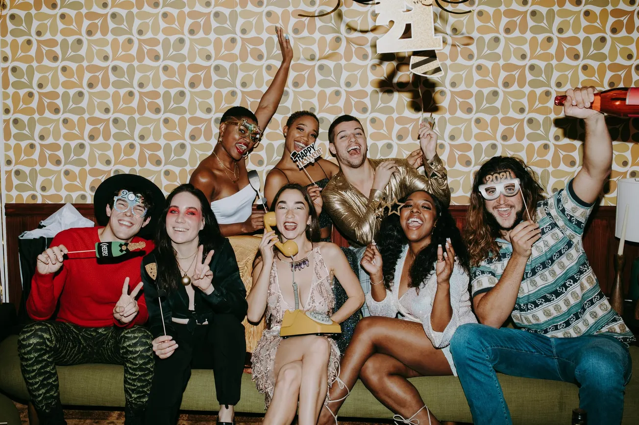 A joyful group of eight friends celebrating indoors with party props and fun attire, against a retro-patterned wall, conveying excitement and togetherness.