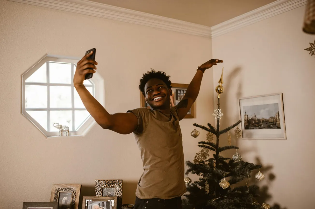 A person joyfully takes a selfie near a decorated Christmas tree, smiling widely. The room is cozy with framed photos and soft lighting, evoking warmth.