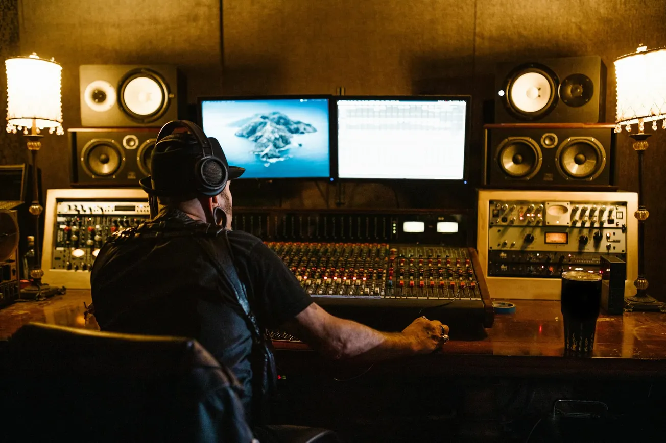 A person wearing headphones sits at a dimly lit recording studio console with multiple screens and speakers. The atmosphere is focused and creative.