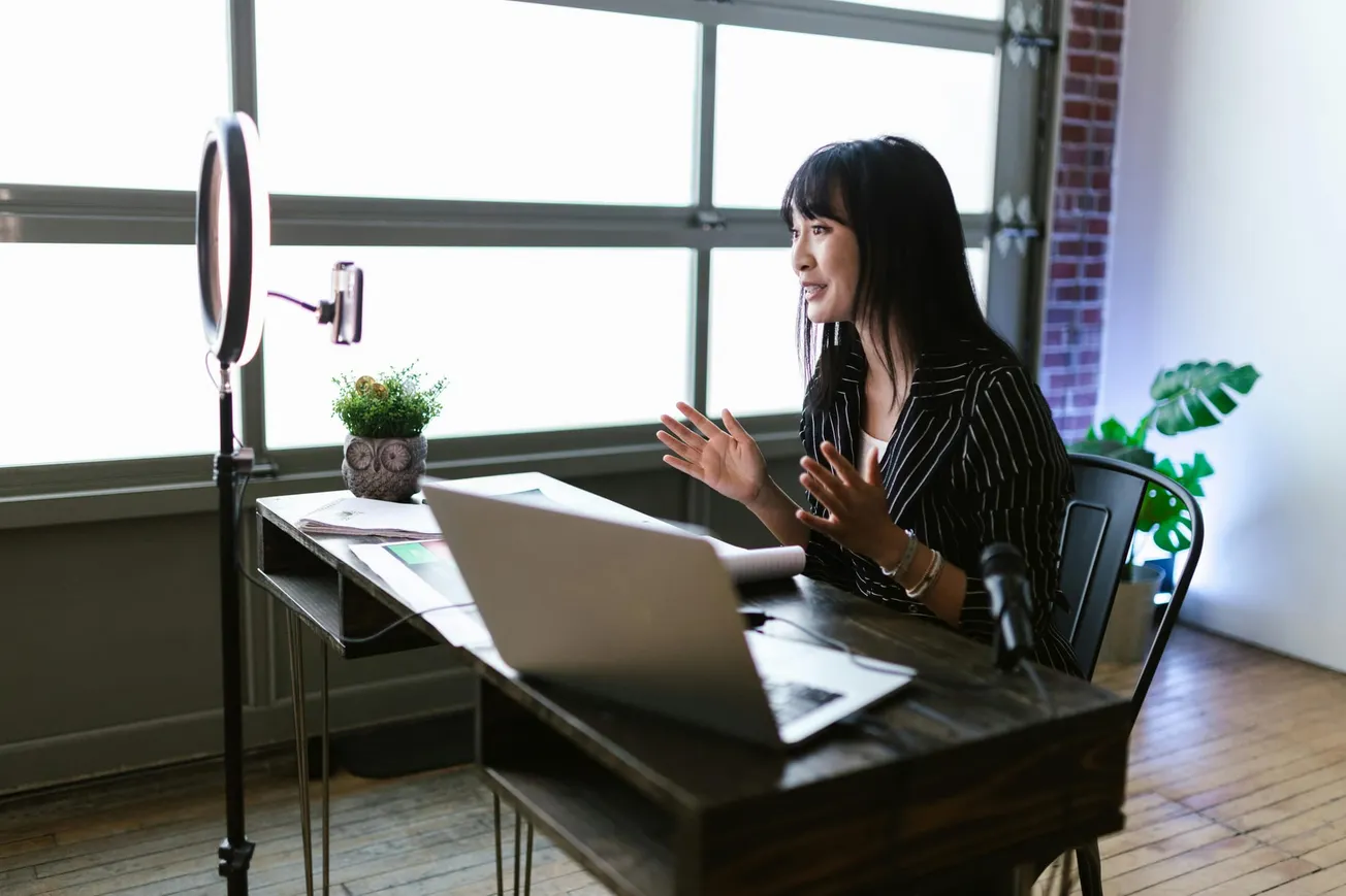 A woman in a striped suit sits at a desk in a bright room, speaking to a camera with a ring light. She's recording with a laptop and microphone, appearing engaged and animated.