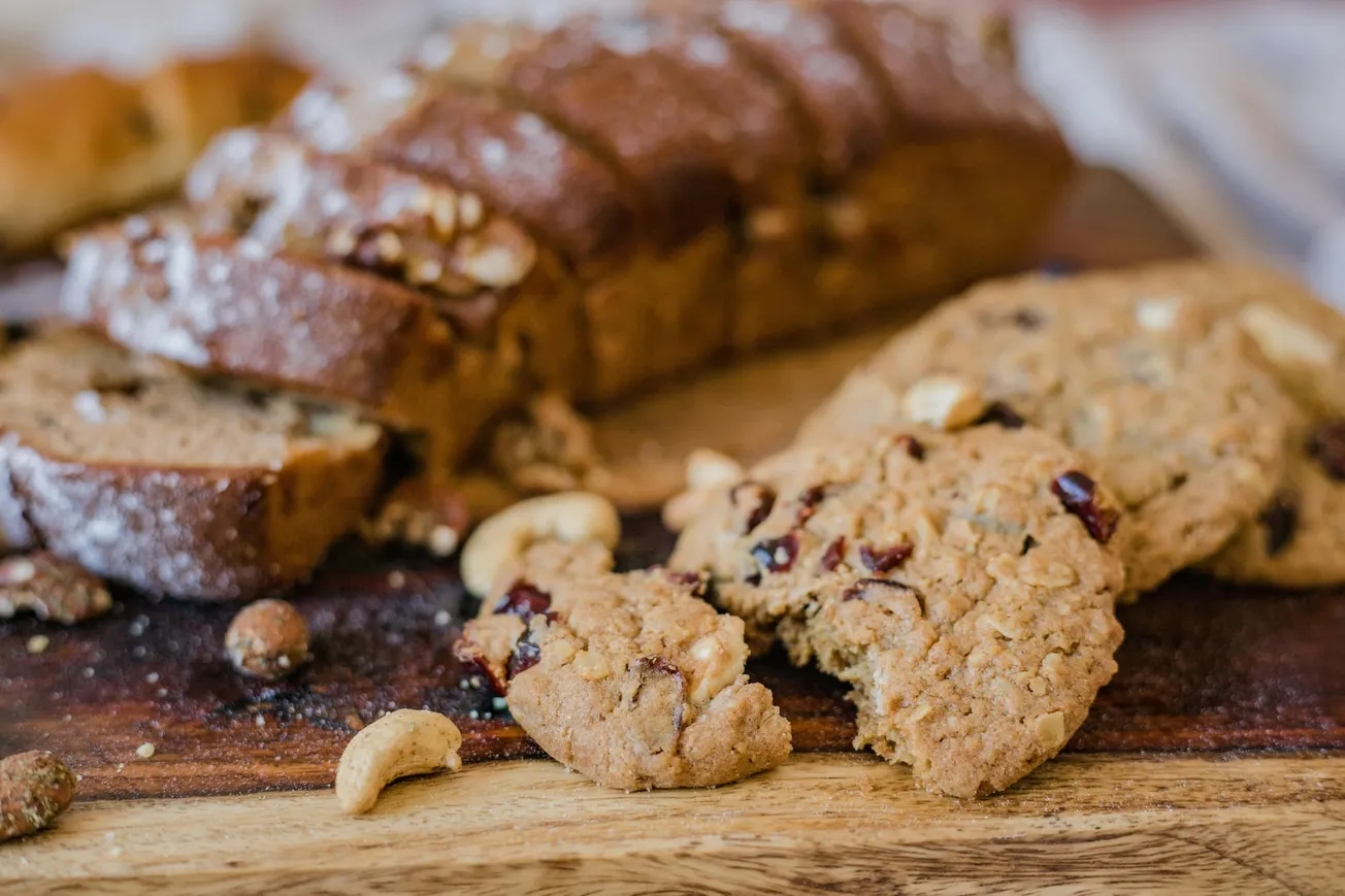 Close-up of sliced nut bread and oat cookies with nuts and dried berries on a wooden board. The scene conveys a warm, homemade feel.