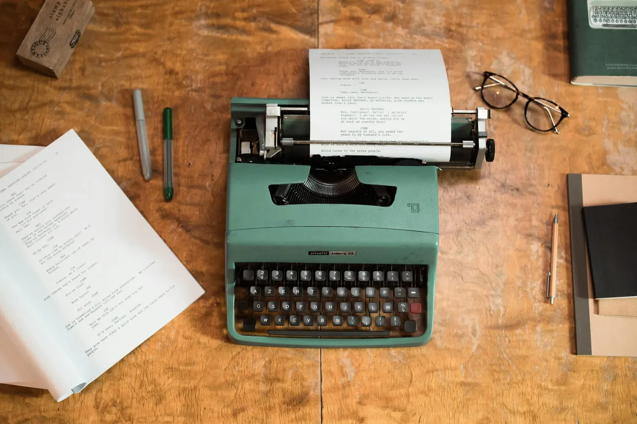 A vintage green typewriter with paper sits on a wooden table, surrounded by a script, pens, a pair of glasses, and a notebook, evoking a nostalgic writing scene.