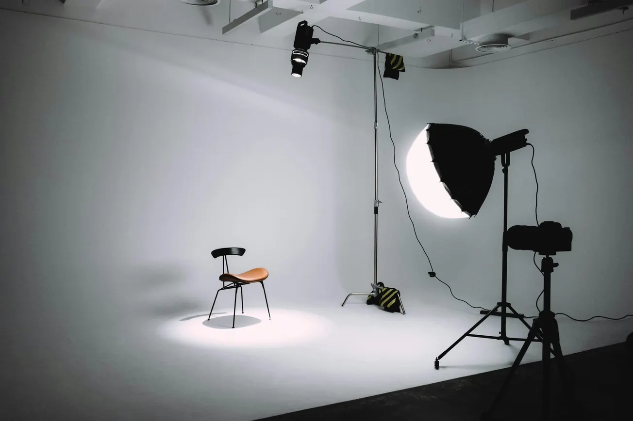 Photography studio setup with an empty chair spotlighted against a white backdrop. Surrounding equipment includes a tripod, lights, and a large softbox.
