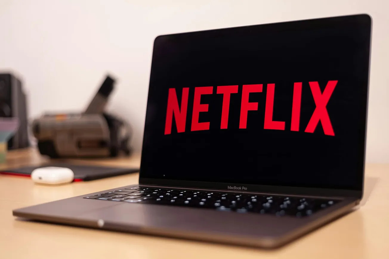 A MacBook Pro on a desk displays the Netflix logo on its screen. Nearby are an AirPods case and a blurred background with office items, suggesting a relaxed mood.