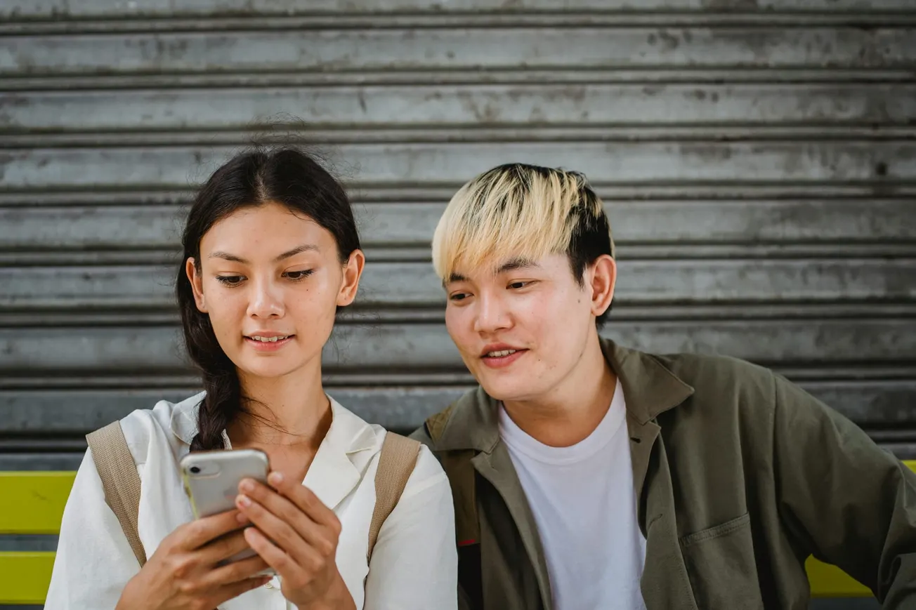 Two people sit on a bench, smiling while looking at a smartphone. They're in front of a metal shutter. The mood is casual and friendly.