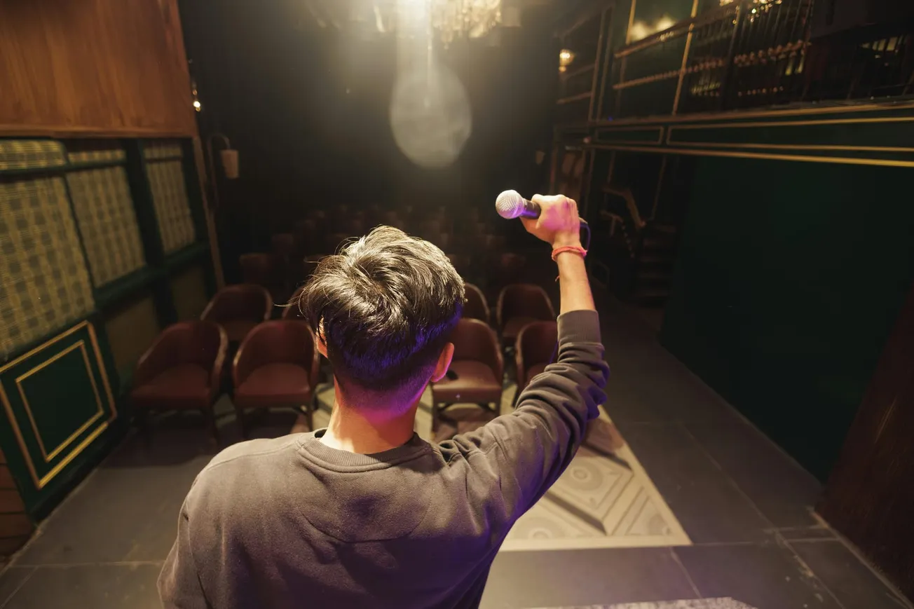 A person holding a microphone stands on a stage facing an empty audience in a dimly lit theater. The setting feels intimate and anticipatory.