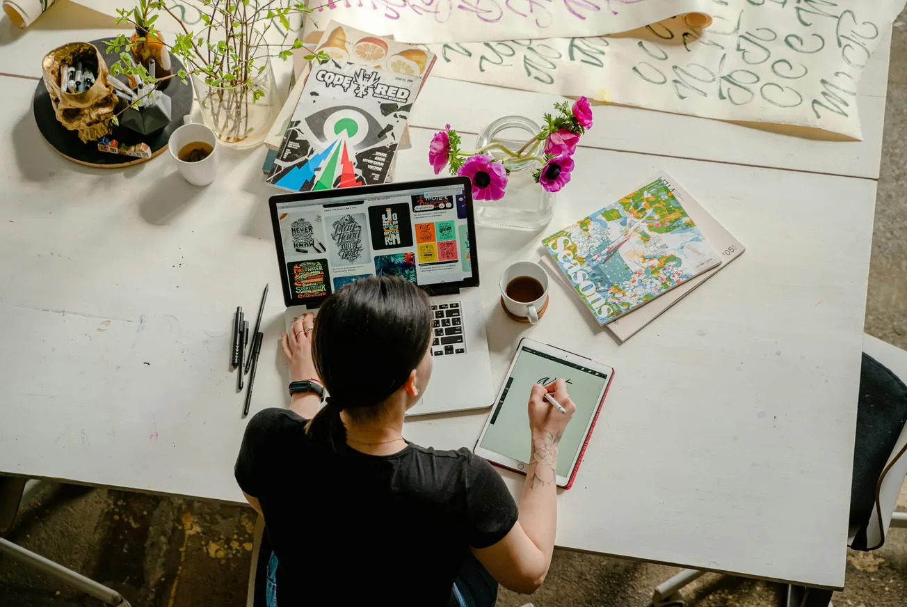 A woman sits at a white table, sketching on a tablet, surrounded by art supplies, flowers, coffee, and a laptop displaying colorful designs.