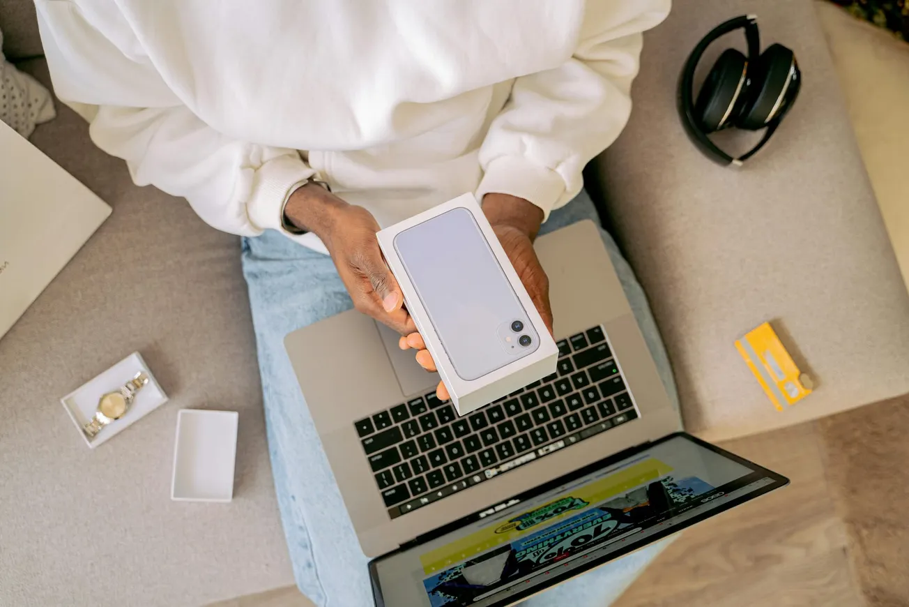 A person in a white sweater holds a smartphone box, seated on a sofa with a laptop. Nearby are headphones and a credit card, suggesting excitement and anticipation.