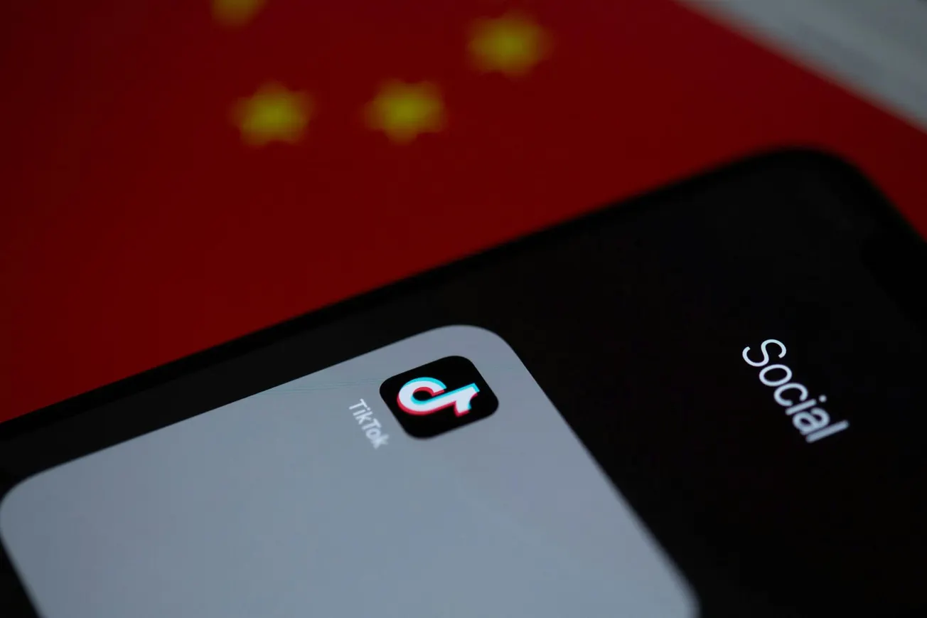 Close-up of a smartphone screen showing the TikTok app icon under a folder labeled "Social." In the background, the red flag of China is partially visible.