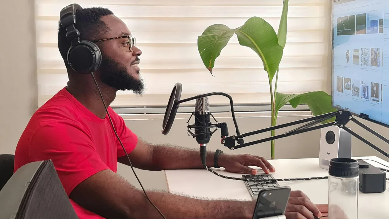 A man in a red shirt and headphones sits at a desk with a microphone, working on a computer. A large plant is in the background, conveying a relaxed home office vibe.