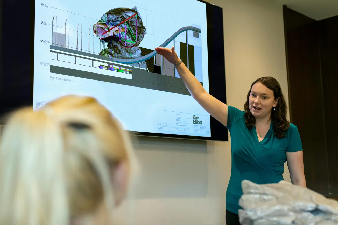 A woman in a teal shirt presents in front of a screen displaying technical drawings and graphs. She gestures energetically, engaging an audience.