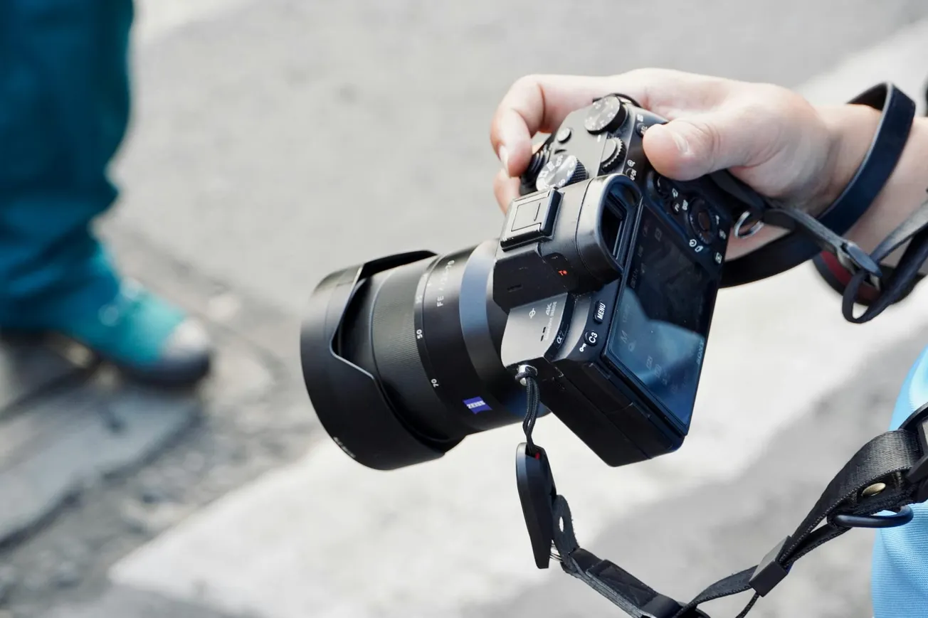 A person holds a digital camera with a large lens, ready to take a photo. The background is a blurred street scene, conveying a sense of focus and anticipation.