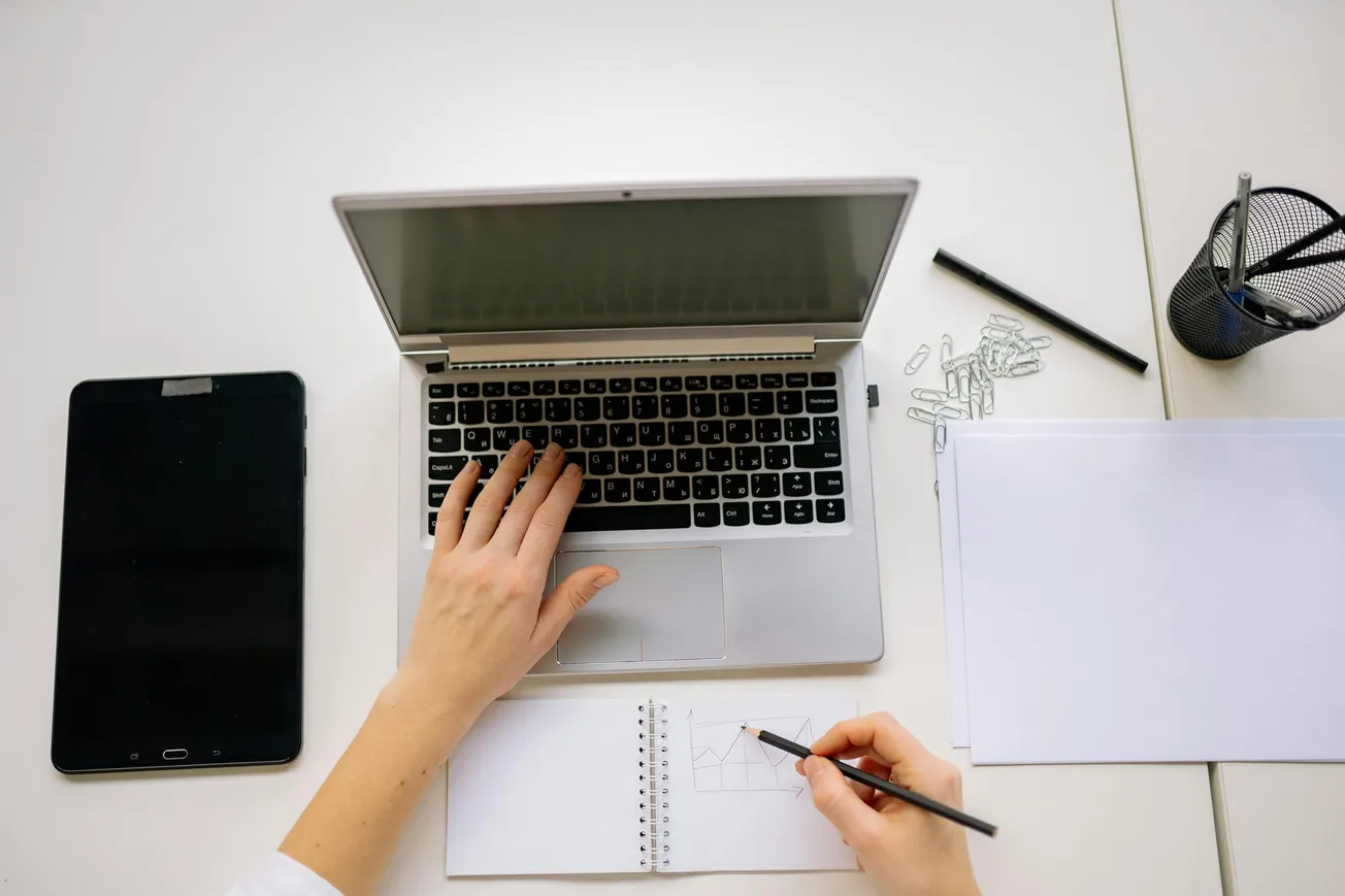 Overhead view of a person working at a desk with a laptop, tablet, notebook, and office supplies, conveying focus and productivity.