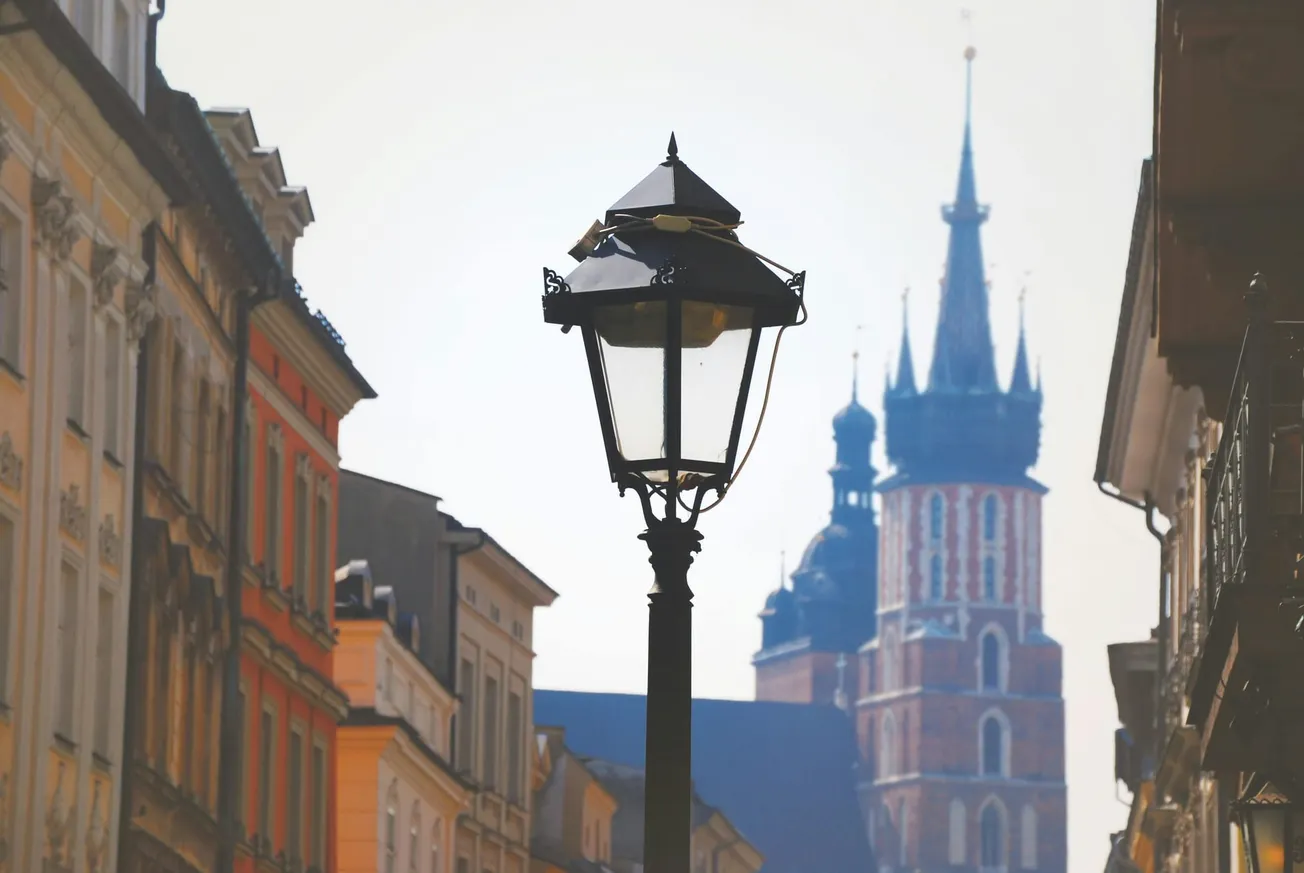 A vintage street lamp in focus with blurred historic buildings, including a church tower, in the background under a clear sky. Urban and timeless ambiance.