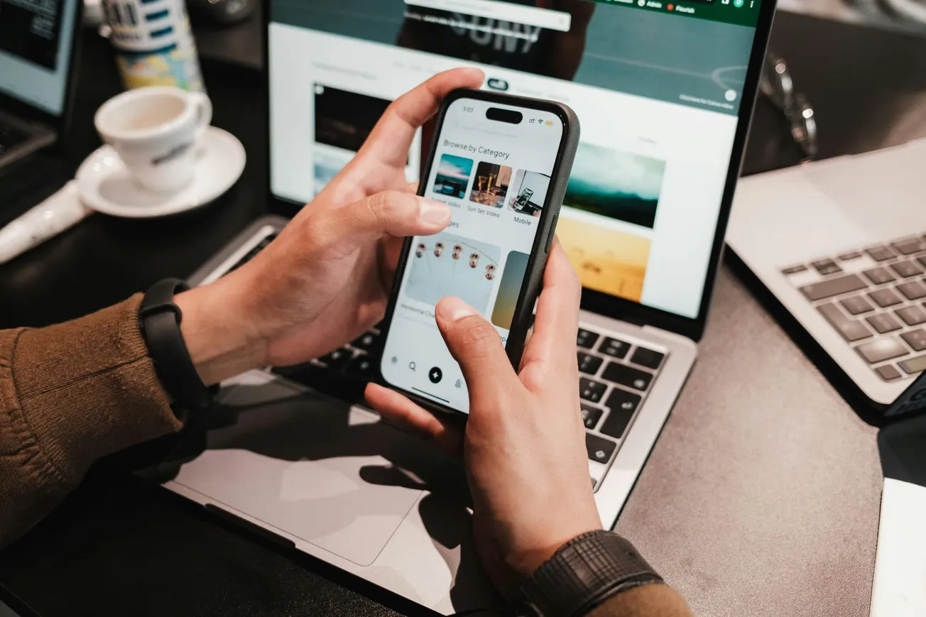 Hands holding a smartphone showing a shopping app, above a laptop with a cup of coffee nearby. The scene conveys a busy, tech-focused environment.