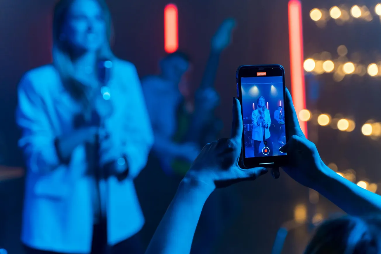 A person holds a phone recording a live music performance on stage. The scene is softly lit with blue tones and red neon lights, creating an energetic vibe.