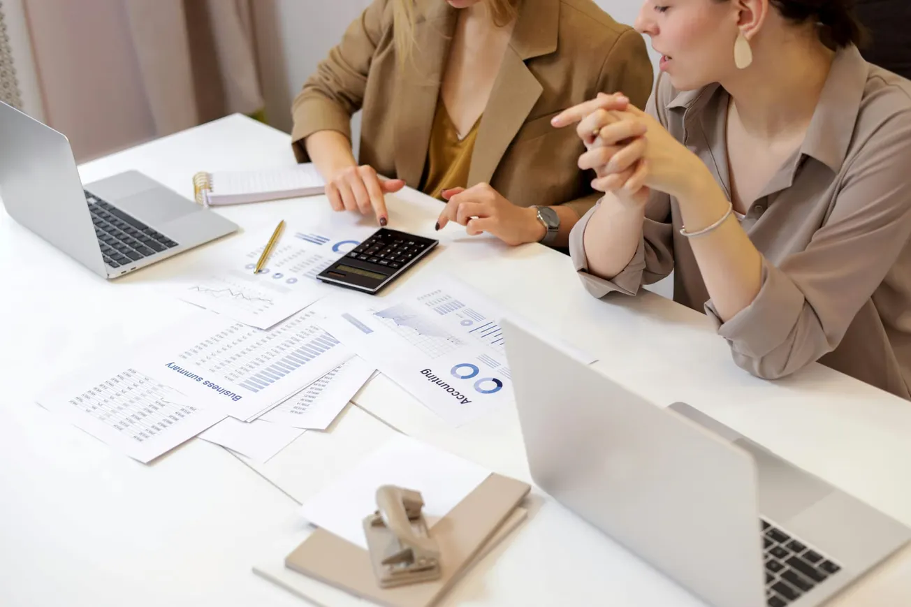 Two people collaborate at a desk with laptops, charts, and a calculator. They discuss financial documents, creating a focused, professional atmosphere.