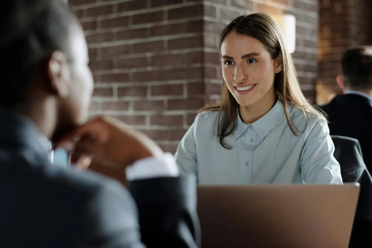 A woman in a light blue shirt smiles while sitting at a table with a laptop, engaging in a conversation with a blurred figure in the foreground.