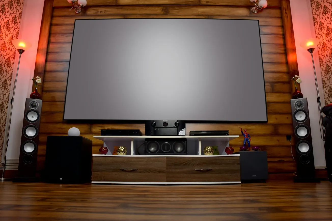 Home theater room with a large screen, wooden wall, and floor. Two tall speakers and red-lit lamps flank the setup. Elegant and cozy ambiance.