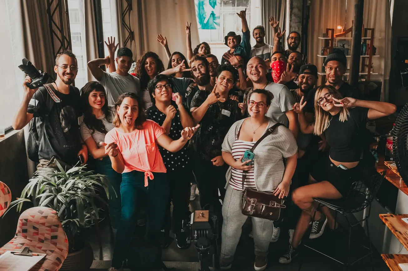 A joyful group of diverse people posing indoors, smiling and gesturing peace signs. The setting is casual, with natural light from large windows.