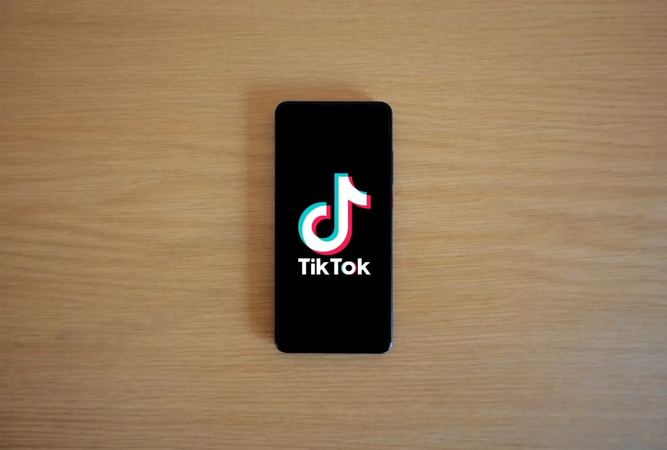 A smartphone displaying the TikTok logo, set on a light wooden surface. The black screen highlights the colorful logo, conveying a modern and digital theme.