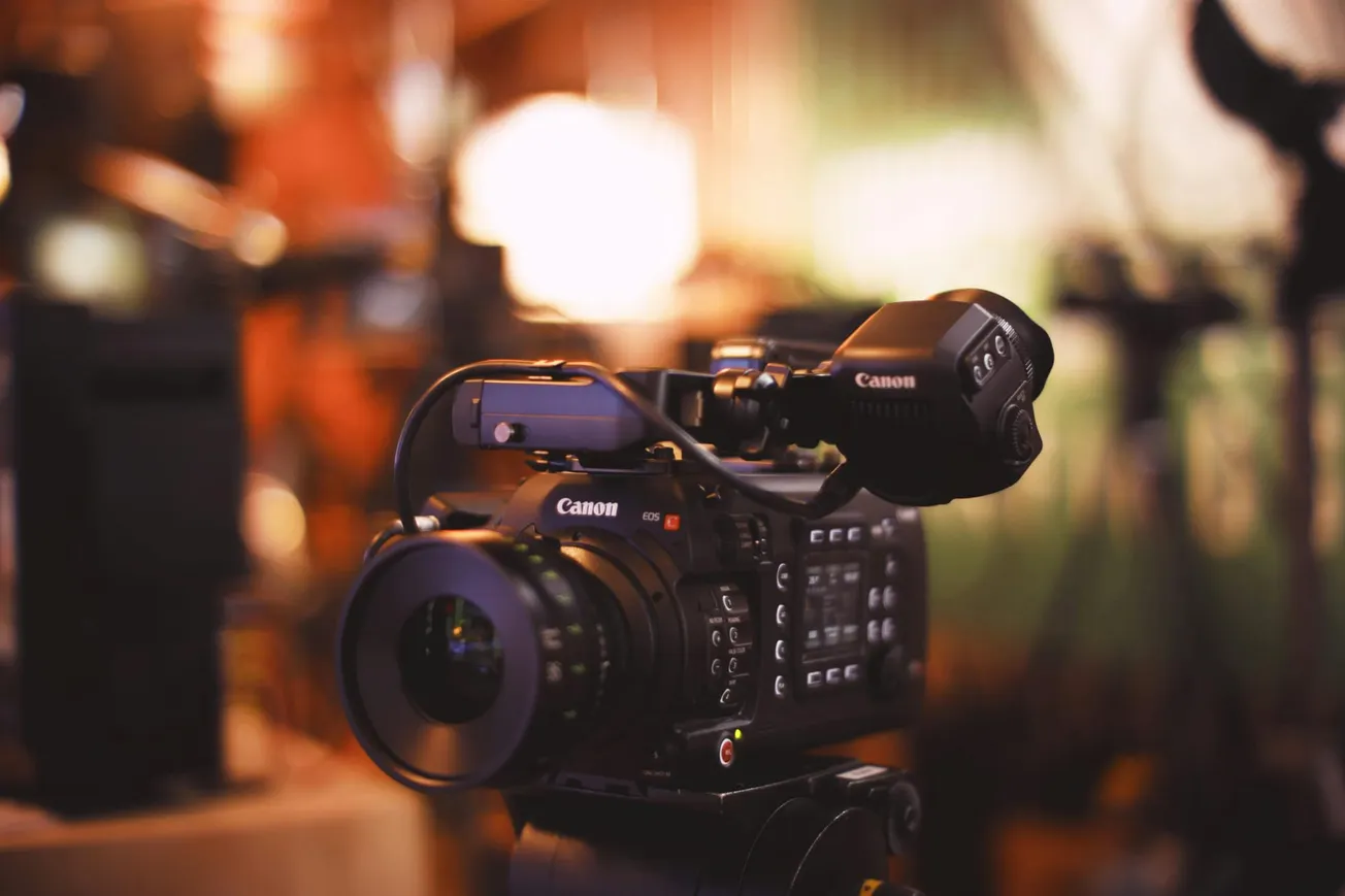 Close-up of a professional Canon video camera set up in a warmly lit studio. The background is blurred, highlighting the camera's intricate features.