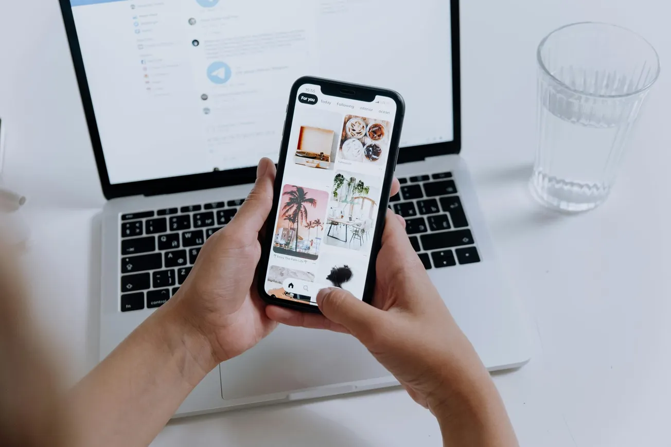 Hands holding a smartphone displaying a photo gallery, in front of an open laptop on a desk. Nearby is a half-full glass of water, suggesting a work setup.