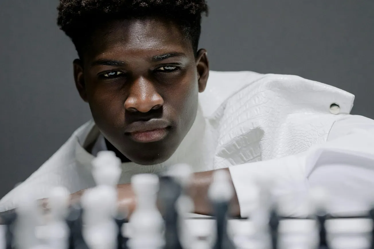 A focused individual in a white jacket leans over a chessboard, intently observing the pieces. The scene conveys concentration and strategy.