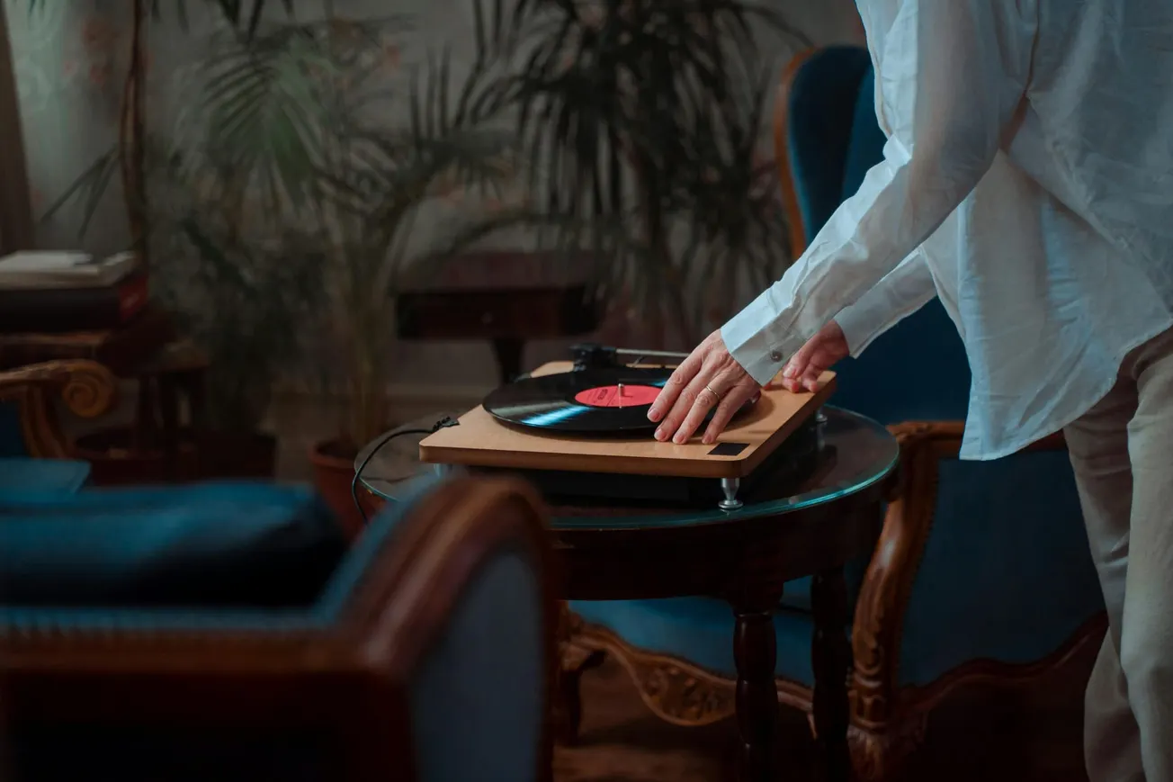 A person in a white shirt places a vinyl record on a turntable, set on a wooden table. The room has vintage furniture and plants, creating a nostalgic ambiance.
