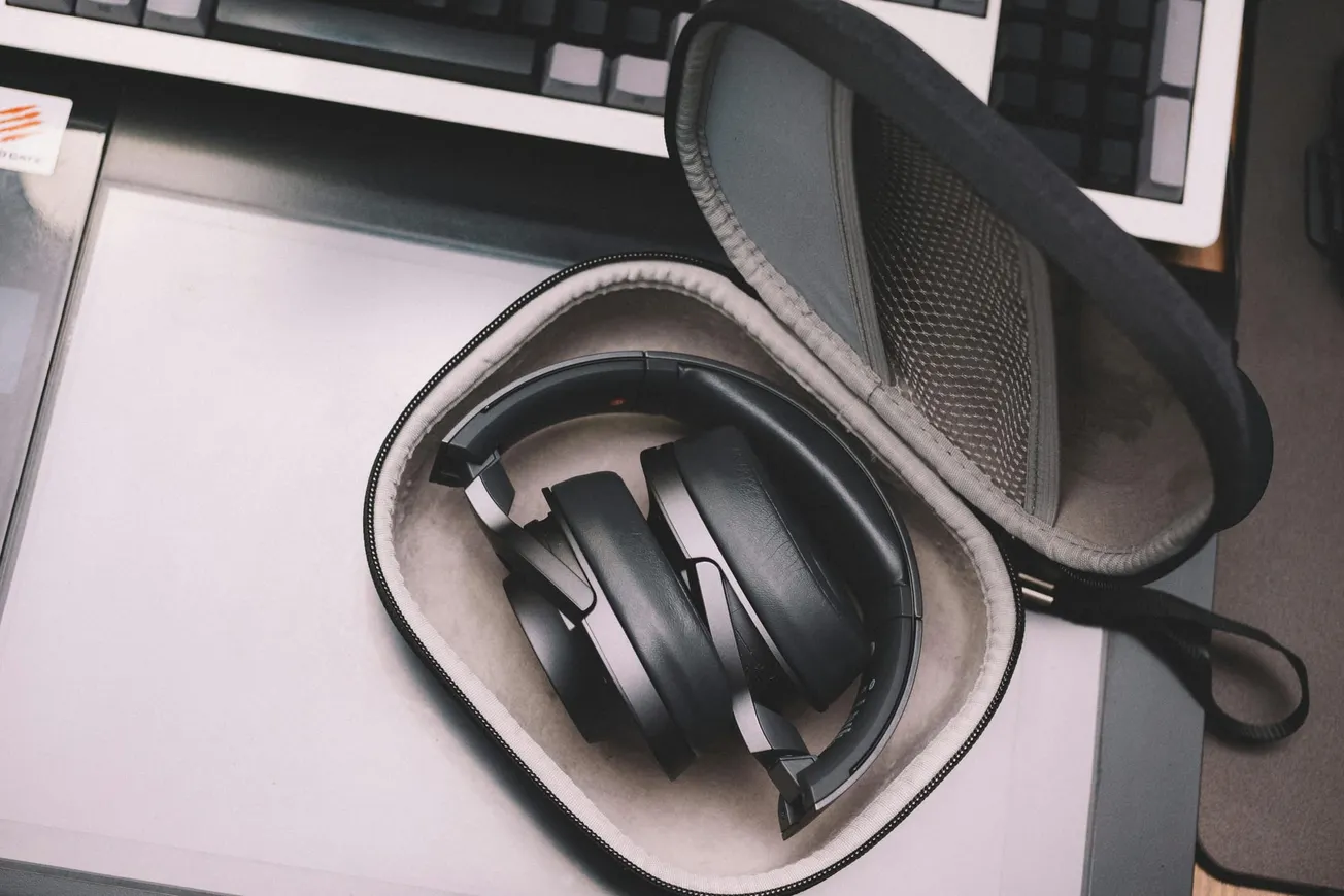 Black over-ear headphones sit neatly in an open, padded carrying case on a desk beside a keyboard, conveying a sense of organization and readiness.