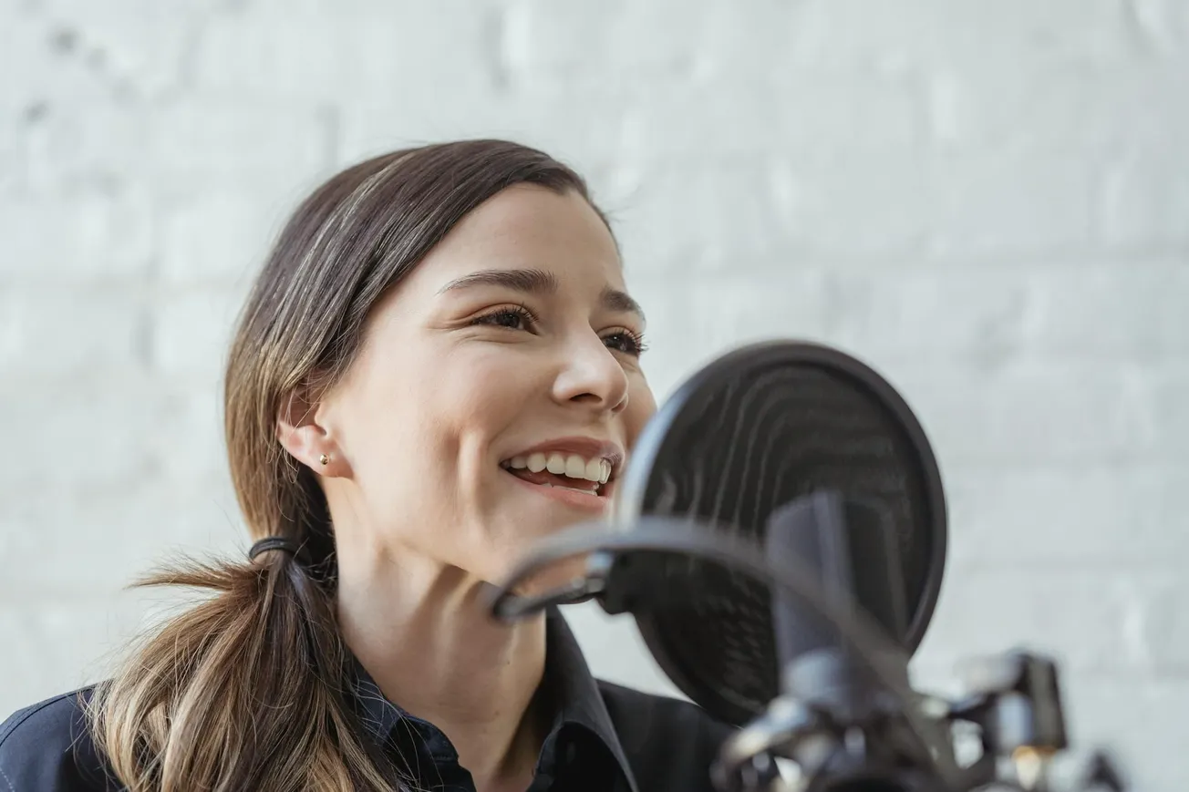 A woman with long brown hair smiles while speaking into a microphone with a pop filter. She appears joyful and engaged, set against a light brick wall.