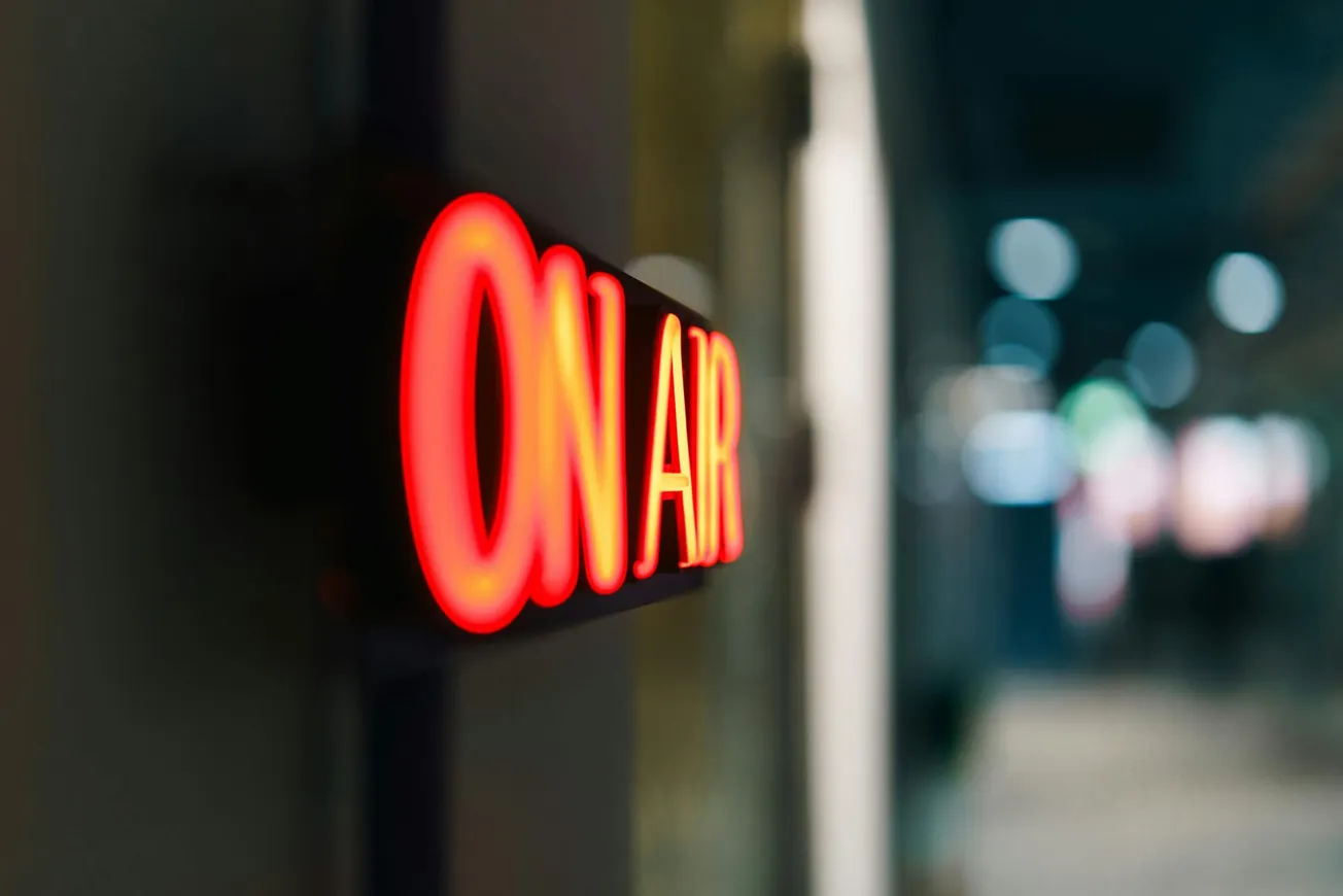 Neon "ON AIR" sign glows in red with a blurred hallway background, conveying a professional broadcast setting and a sense of urgency.