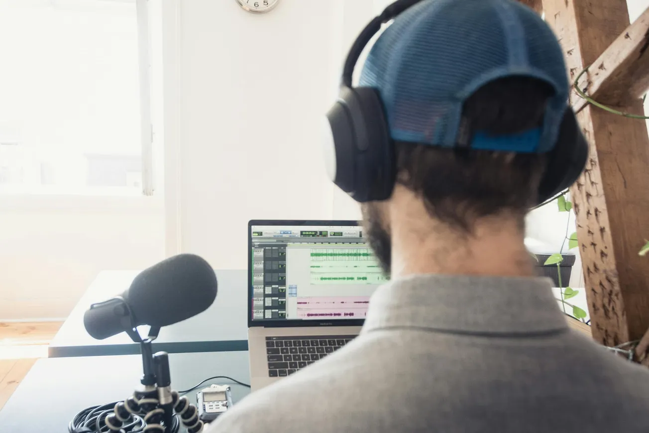 A person wearing headphones sits at a desk with a microphone and open laptop displaying audio software, suggesting a podcast recording setup.