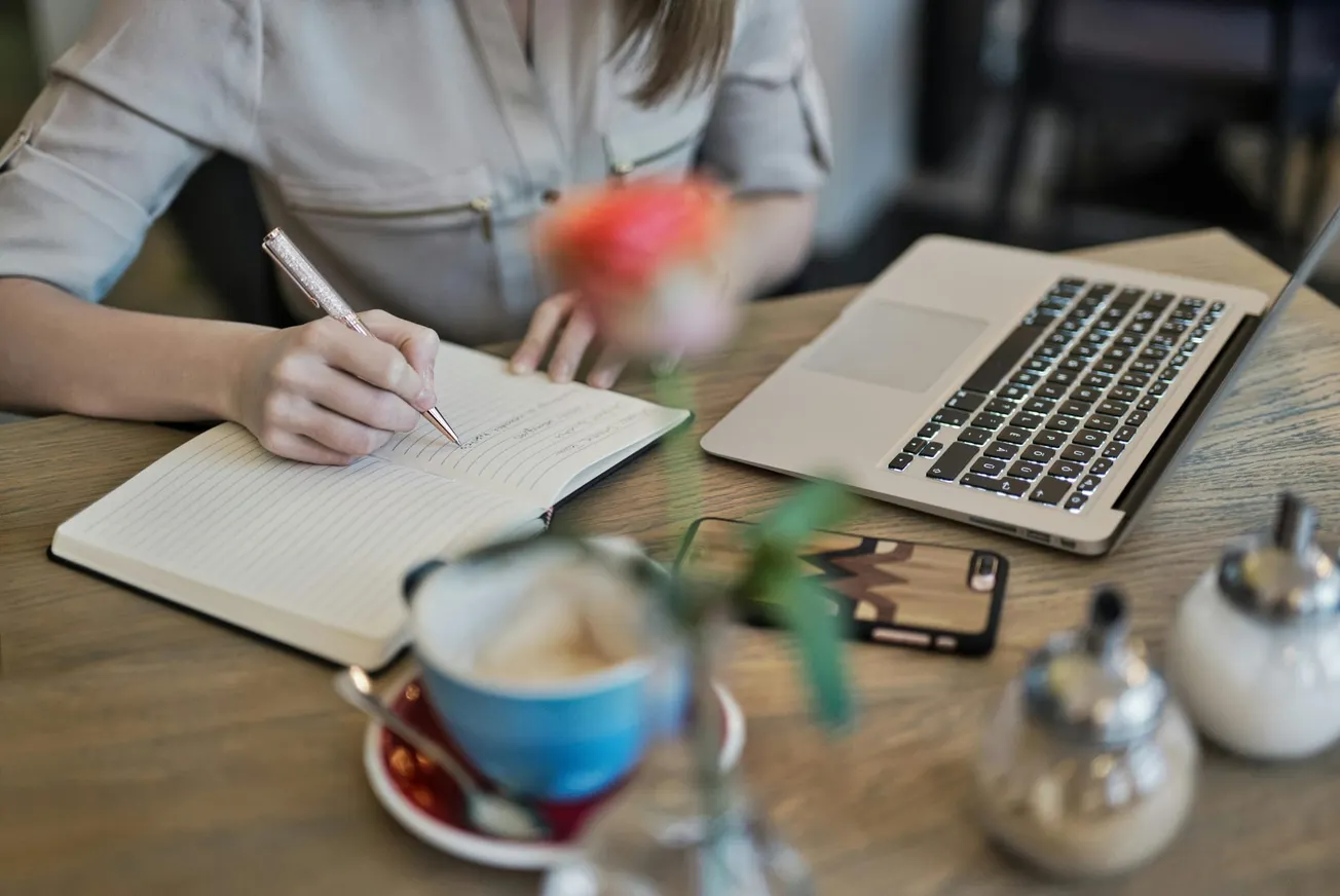 A person writes in a notebook at a wooden table with a laptop, smartphone, and blurred flowers. The scene is calm, suggesting focus and productivity.