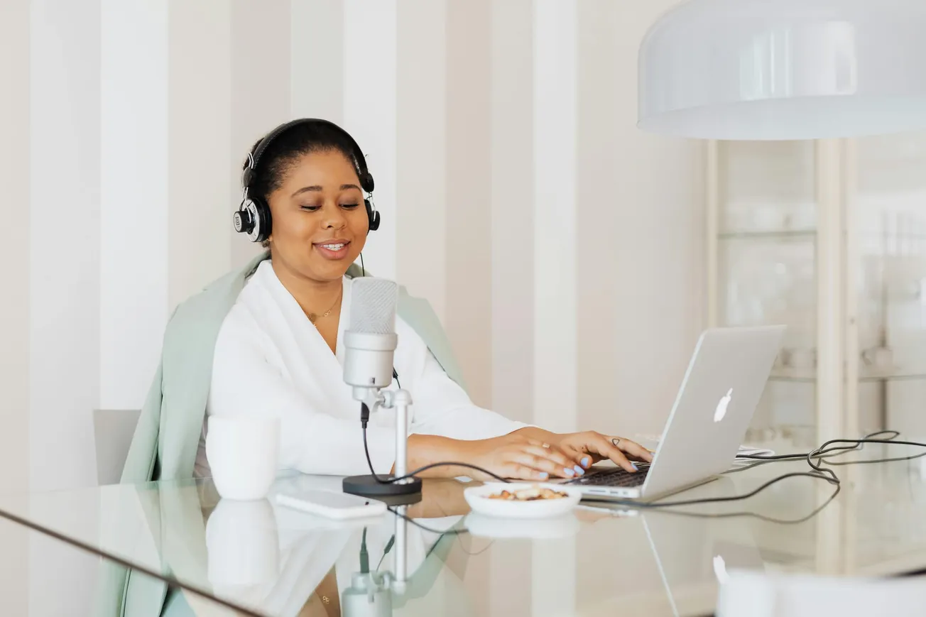 A woman wearing headphones smiles while using a laptop and microphone at a glass table. A mug and snack bowl are nearby, creating a cozy, productive vibe.