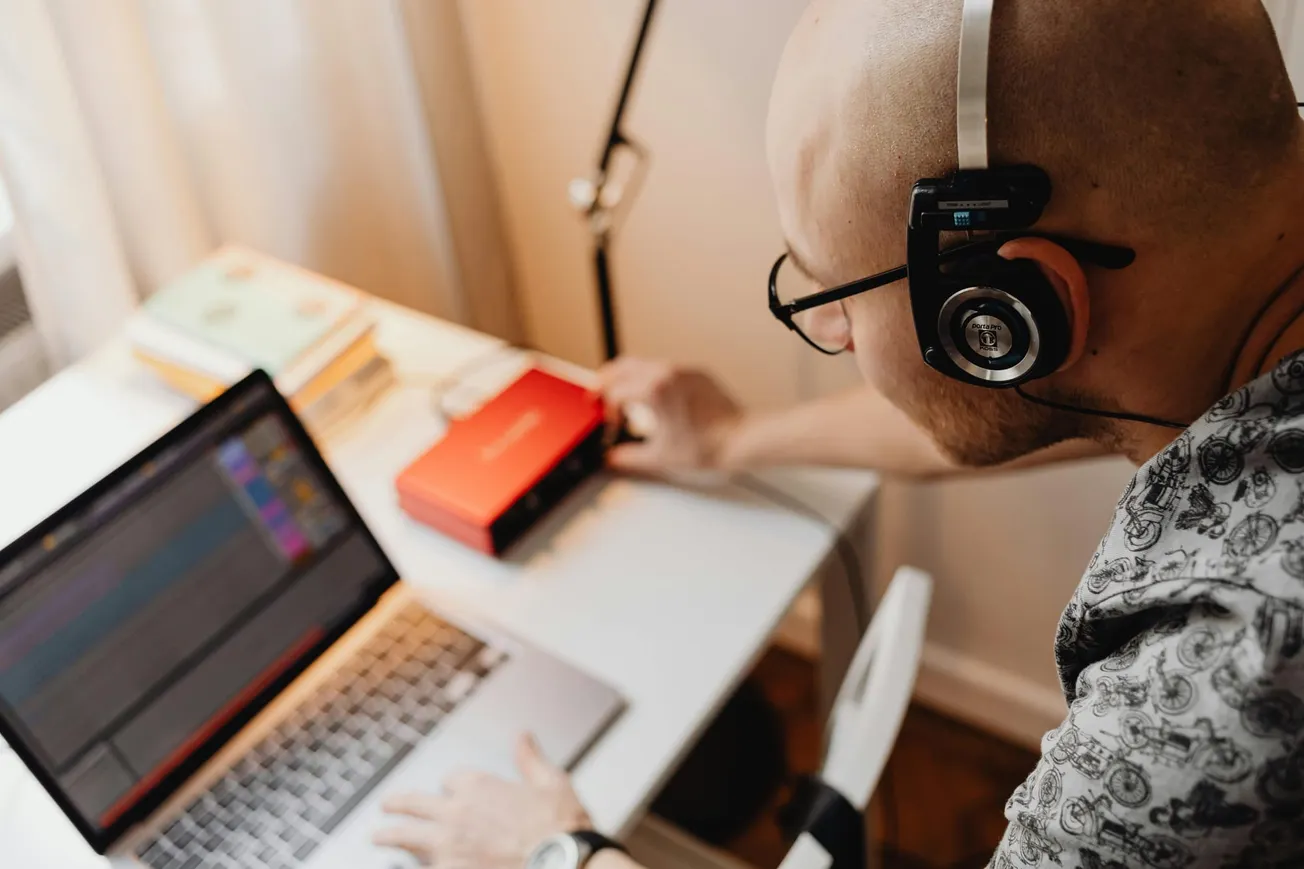 A bald person wearing headphones works on a laptop at a desk, with a red audio interface and books nearby. Focused, creative atmosphere.