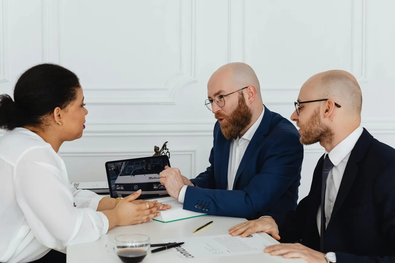 Three professionals in an office setting are engaged in a serious discussion. Two men in suits face a woman holding a pen, with papers and a laptop on the table.