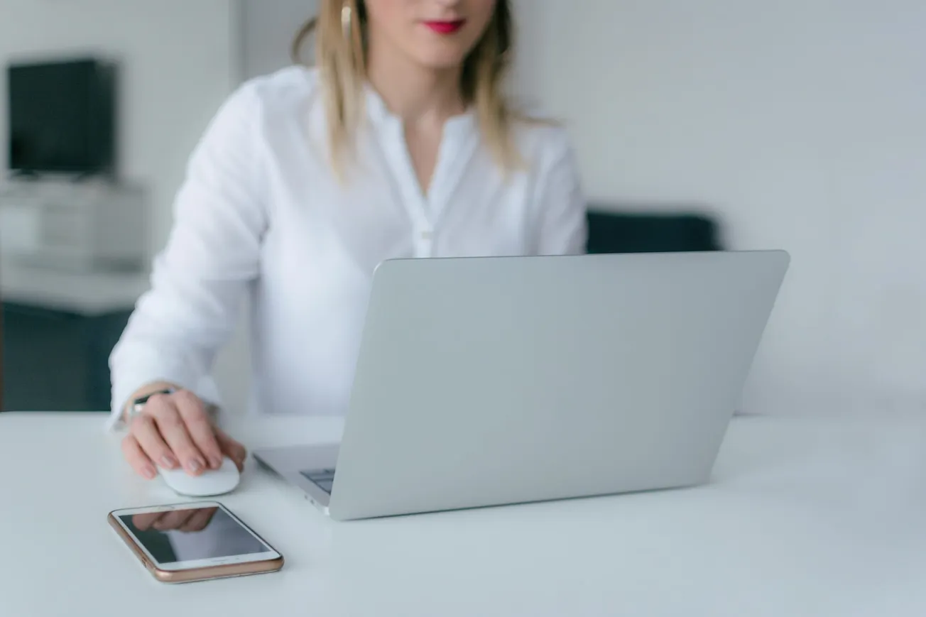 A woman in a white shirt uses a laptop on a white desk, with a smartphone nearby. The setting is a modern, minimalistic office, conveying focus.