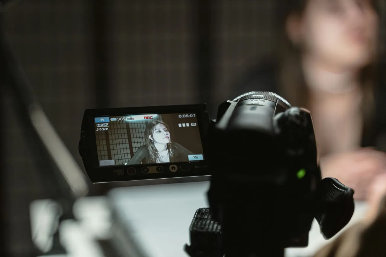 A video camera displays a recording of a woman with long hair, wearing a dark top, in focus. The camera's screen is clear, while she is blurred in the background.