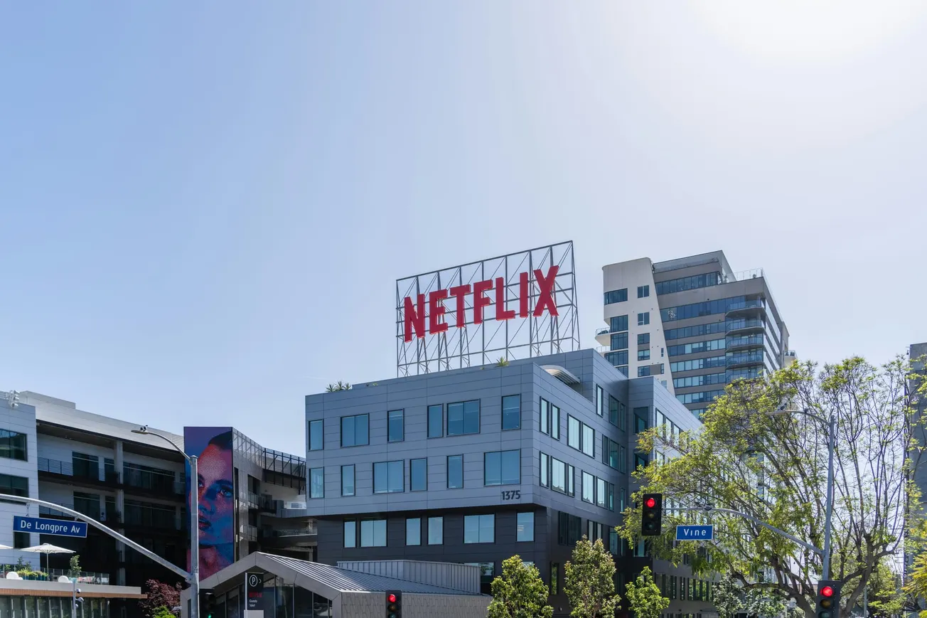 A modern building with a large red Netflix sign on its roof, under a clear blue sky. Trees and road signs are visible in the foreground, creating an urban feel.