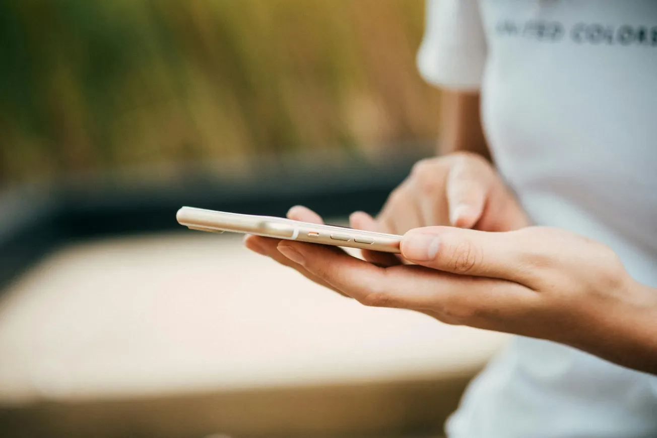 A person in a white shirt is using a smartphone outdoors. Their fingers are tapping the screen, conveying focus and engagement. Background is blurred.