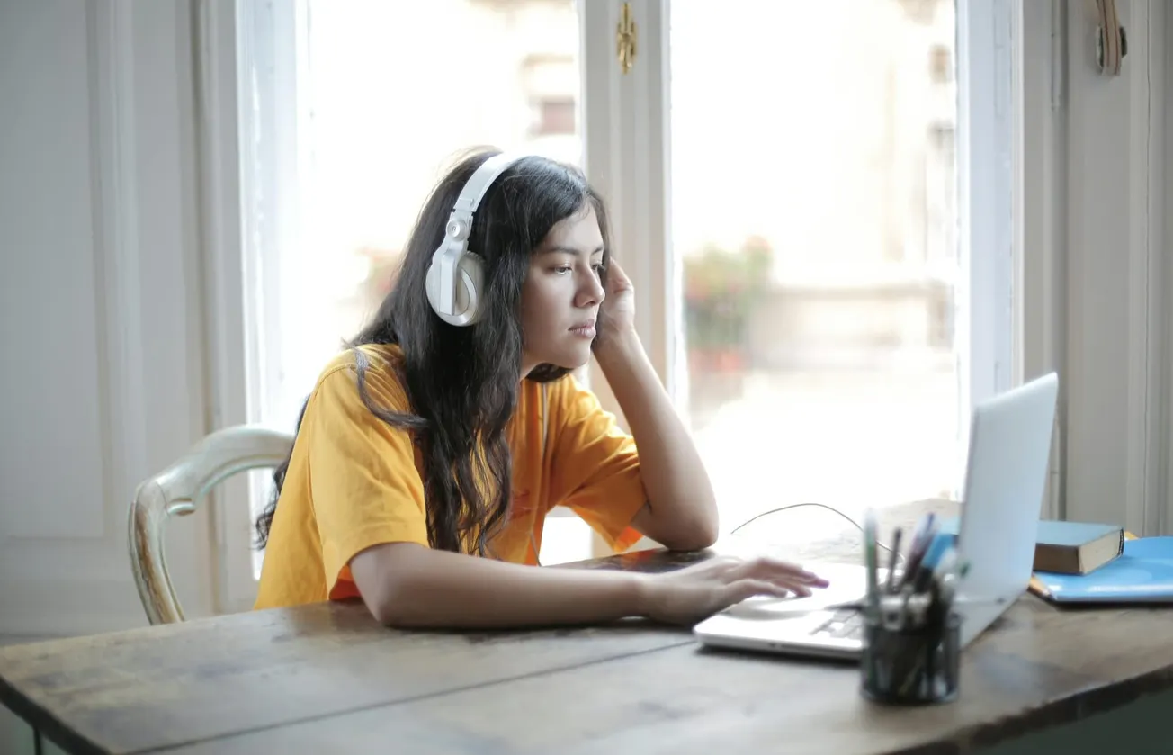 A young woman in a yellow shirt sits at a wooden table, wearing headphones and focusing on a laptop. She appears thoughtful, with light streaming through a large window.