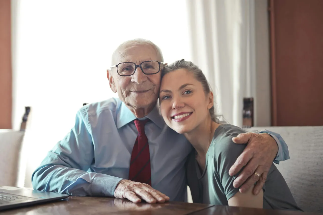 Elderly man with glasses and a blue shirt joyfully embraces a smiling young woman in a gray top. They are seated at a table, conveying warmth and affection.