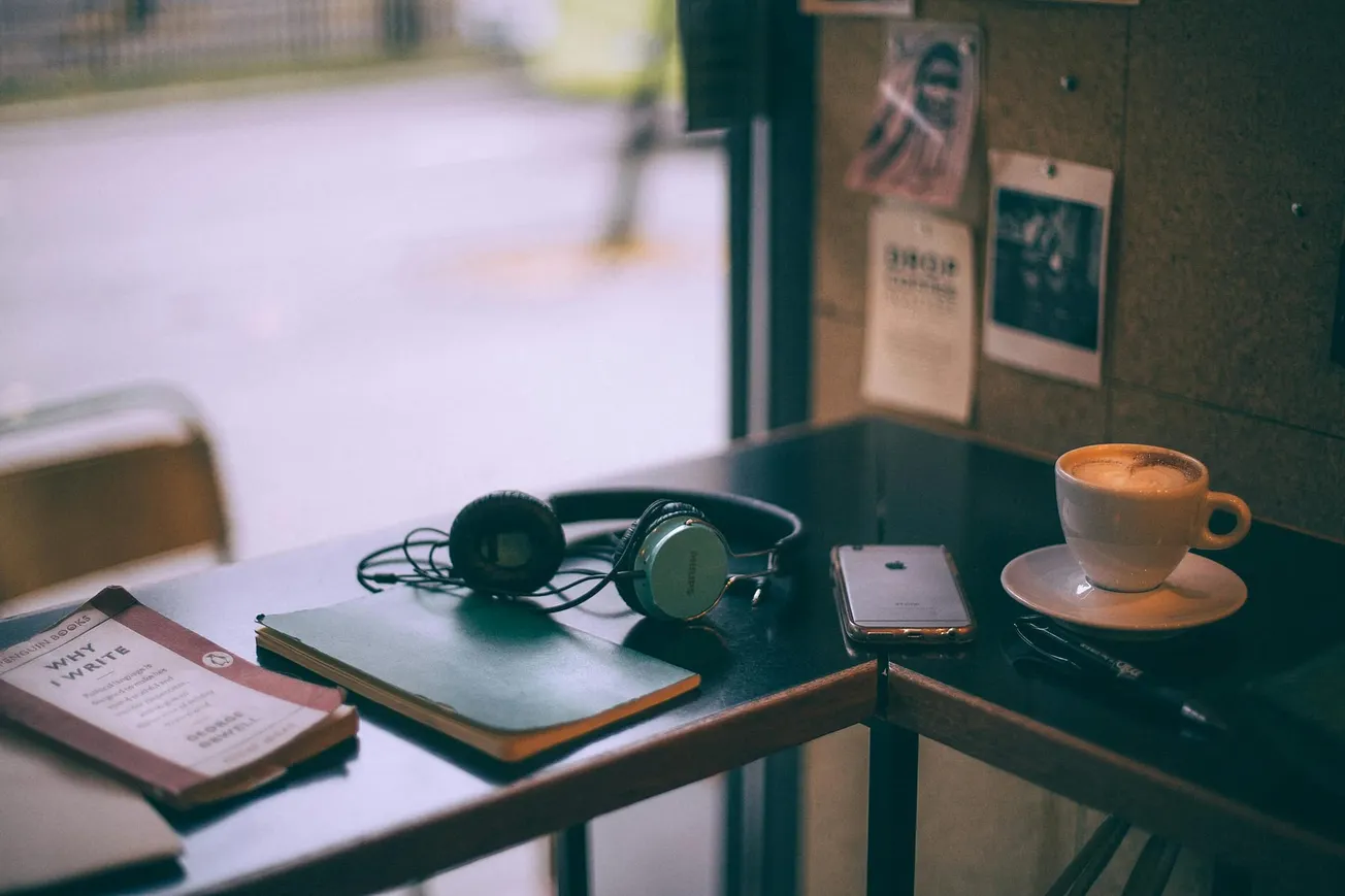 Cozy cafe table with a book, notebook, headphones, smartphone, and a cappuccino. Soft lighting and warm tones create a relaxed, inviting atmosphere.