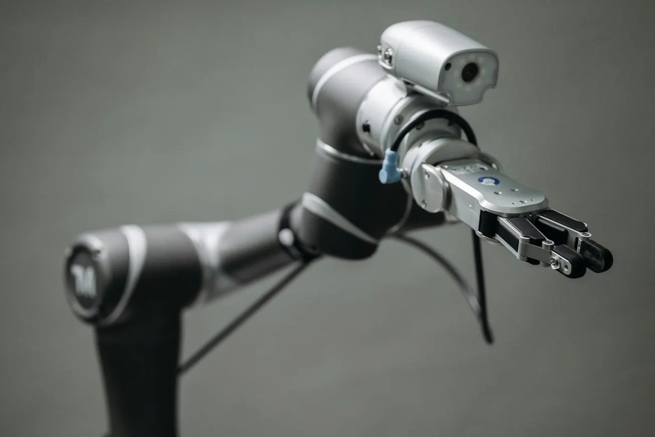 A sleek robotic arm extends forward, showcasing metallic joints and a precise gripper, set against a neutral gray background, conveying technological advancement.