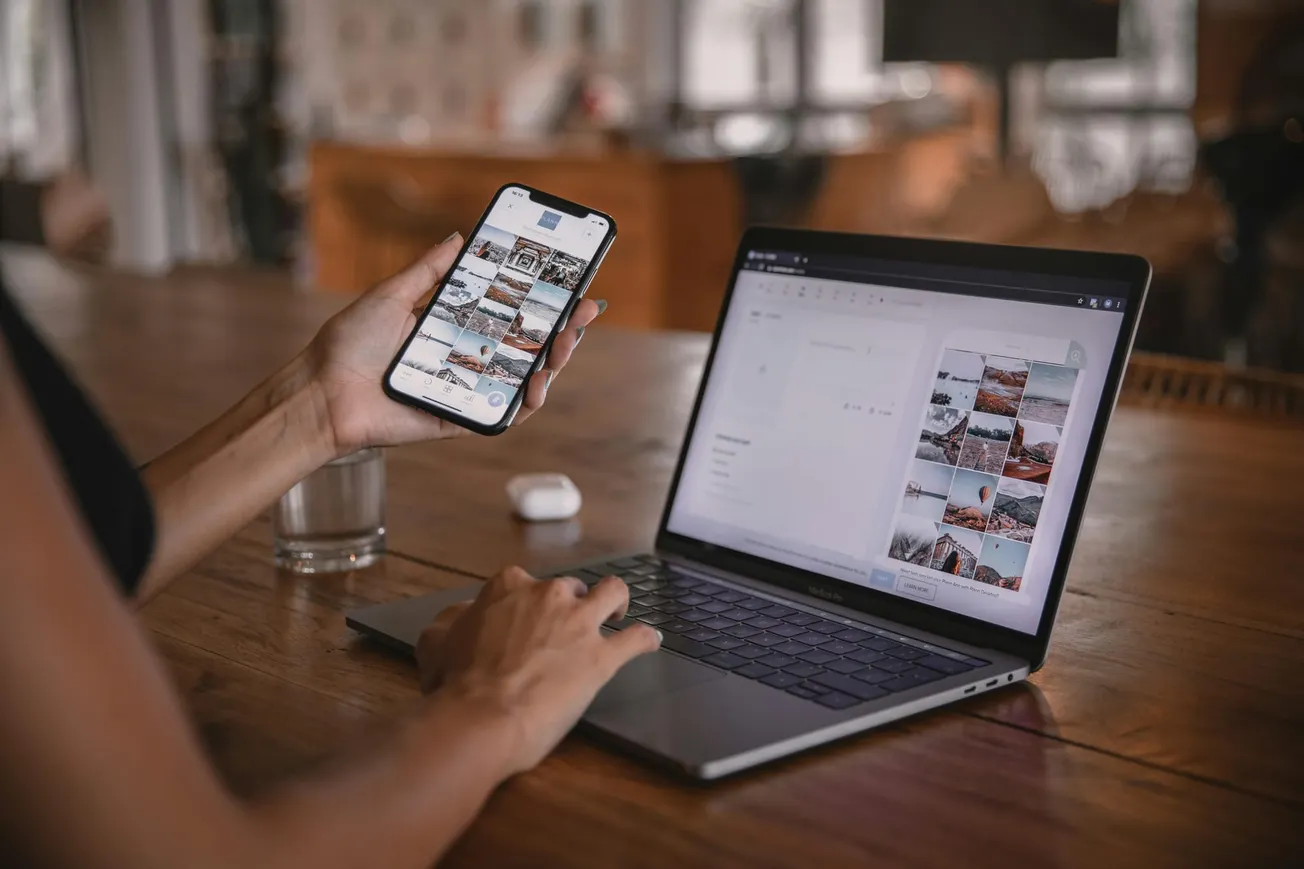 A person sits at a wooden table using a smartphone and a laptop displaying photo editing software. The setting is warm and focused on digital work.