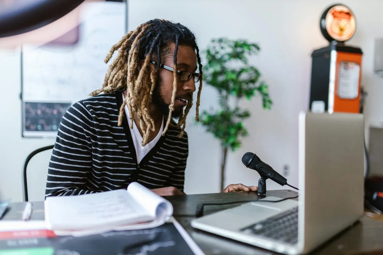 A person with dreadlocks and glasses is speaking into a microphone at a desk with a laptop. Papers are scattered, and a plant is in the background. The room has a focused, creative vibe.