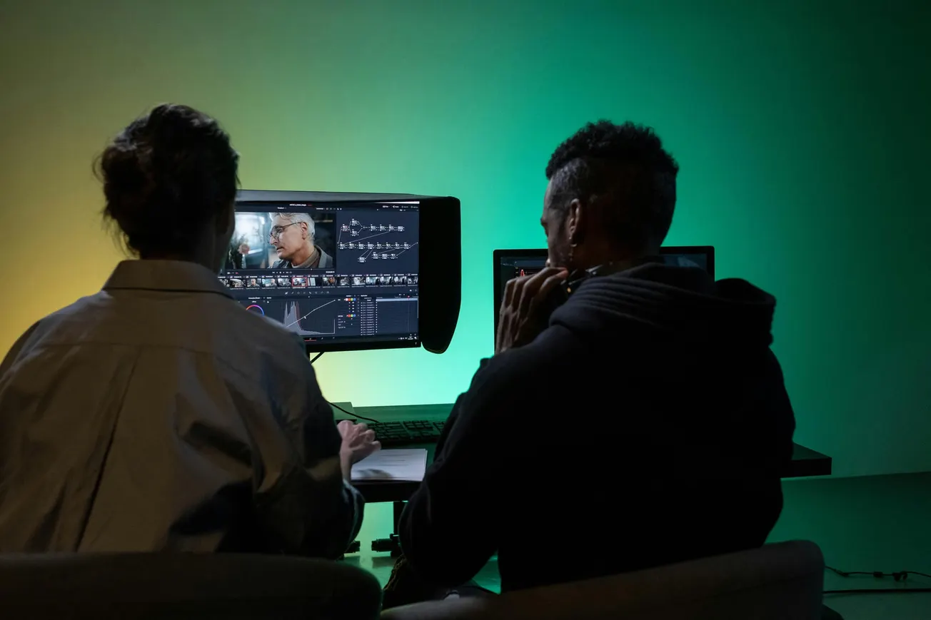 Two people sit in front of a computer monitor displaying video editing software, cast in green and yellow light. The scene conveys focus and creativity.