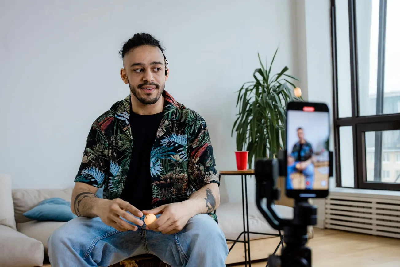 A man with a tropical shirt and jeans sits in a cozy room, smiling at a smartphone on a tripod. A plant, couch, and red cup are in the background. Relaxed atmosphere.